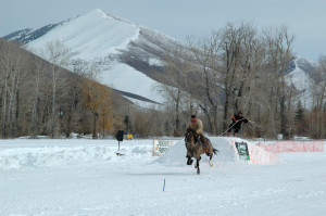 ski joring horse and horseback rider pull skier over jump on course with snowy mountain in background